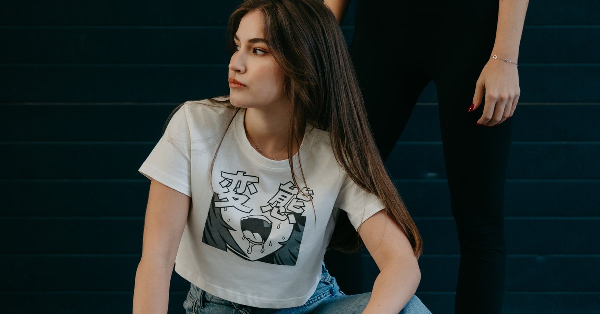Dvě modelky v anime streetwearu Yugymo — klečící dívka v krémovém crop topu s japonským potiskem před tmavou stěnou